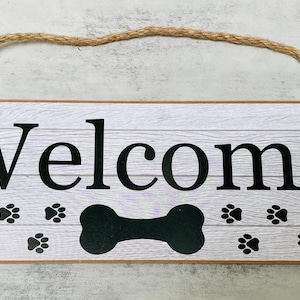 May include: A white wooden sign with the word "Welcome" in black lettering. The sign has a black bone shape and paw prints on it. The sign is hanging from a rope.
