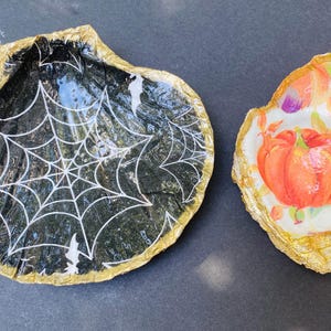 May include: Two decorative seashell dishes. One is black with a white spiderweb design and gold trim. The other is white with an orange pumpkin design and gold trim. Perfect for Halloween or fall decor.