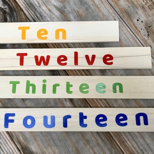 May include: Four wooden blocks with the words "Ten", "Twelve", "Thirteen", and "Fourteen" painted on them in different colors. The blocks are arranged in a stack, with the largest block at the bottom and the smallest block at the top.