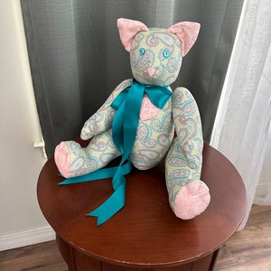 May include: A teal and pink paisley patterned stuffed cat with a teal satin bow. The cat is sitting on a wooden table.