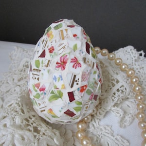 May include: A decorative mosaic egg made from china fragments, adorned with floral and geometric designs in red, pink, yellow, and green. A strand of faux pearls is placed on a white lace surface.