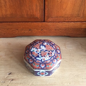 Puede incluir: Caja de alhajas octogonal de porcelana con un diseño floral colorido. La tapa presenta un fondo blanco con flores rojas, azules y verdes. La base de la caja es azul con detalles dorados.