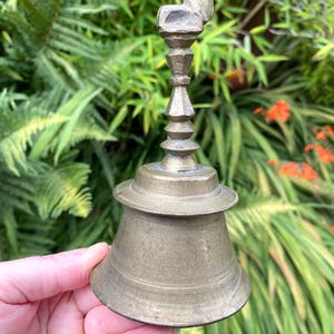 May include: Vintage brass bell featuring a donkey figurine. The bell has a flared base and a detailed, tiered stem. The bell is set against a backdrop of green foliage and orange flowers.