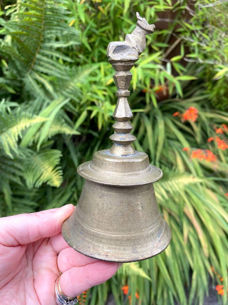 May include: Vintage brass bell featuring a donkey figurine. The bell has a flared base and a detailed, tiered stem. The bell is set against a backdrop of green foliage and orange flowers.