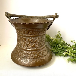 May include: An antique copper bucket with an ornate floral design and a curved handle. The bucket is in good condition with a patina finish.