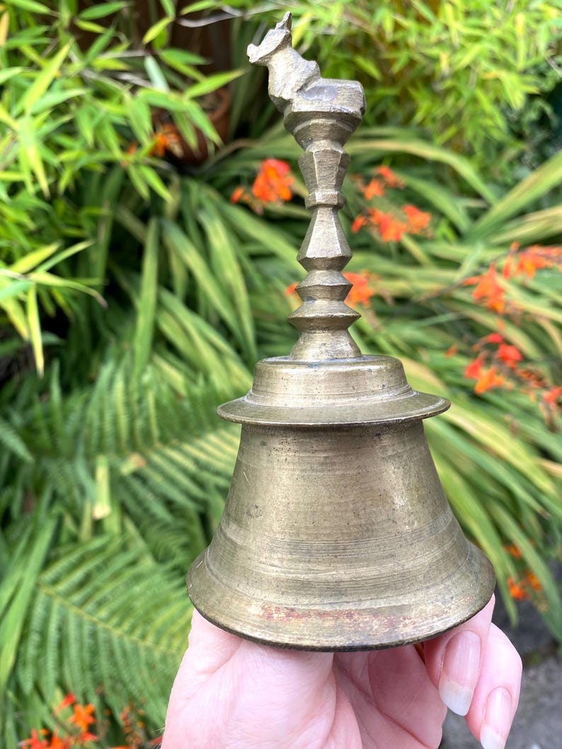 May include: Vintage brass bell with an ornate design. The bell features a flared base and a decorative handle with a small animal figure. The bell has a warm, aged brass colour, set against a backdrop of blurred green foliage.