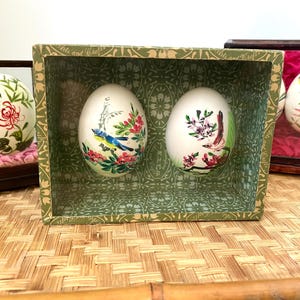 Puede incluir: Huevos pintados decorativos con diseños florales y de aves. Dos huevos se exhiben en una caja verde, mientras que otros están en pequeños expositores de madera. Los huevos presentan obras de arte detalladas con acentos rojos, rosas y azules.
