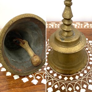 May include: Vintage brass bell with a detailed handle and a worn interior. The bell has a classic shape and a warm, aged patina. The bell is likely made of bronze or a similar alloy.