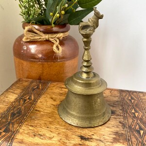 May include: Vintage brass bell with an animal-shaped handle. The bell sits on a wooden surface with a geometric pattern. A brown ceramic vase with greenery is in the background.