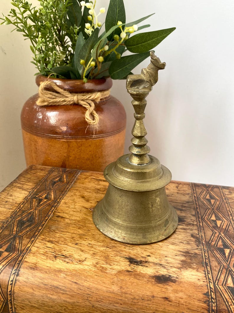May include: Vintage brass bell with an animal-shaped handle. The bell sits on a wooden surface with a geometric pattern. A brown ceramic vase with greenery is in the background.