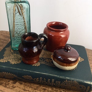以下が含まれることがあります： 緑色の金文字の入った本の上に、茶色の小さな陶器の壺、瓶、蓋付きの壺、そしてボトルが置かれています。