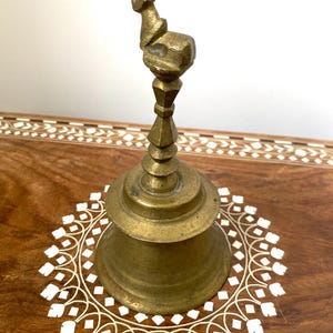 May include: A vintage brass bell with a decorative handle. The bell has a flared base and a detailed top. It is set on a wooden surface with a white floral inlay. The bell is a warm gold colour.
