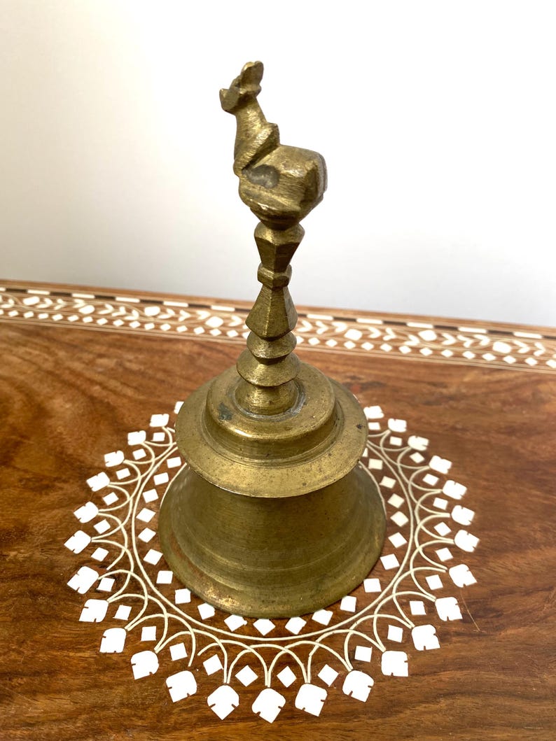 May include: A vintage brass bell with a decorative handle. The bell has a flared base and a detailed top. It is set on a wooden surface with a white floral inlay. The bell is a warm gold colour.