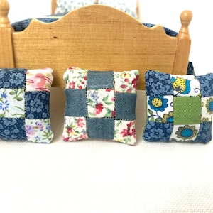 Peut inclure: Trois petits coussins patchwork aux tons de bleu, rose et blanc, avec des motifs floraux et un design carré. Les coussins sont placés devant un cadre de lit en bois, suggérant un décor de maison de poupée ou de chambre miniature.