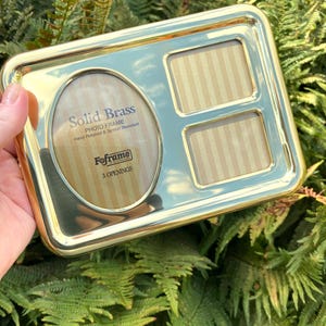 May include: A rectangular solid brass photo frame with three openings. The frame has an oval opening and two smaller rectangular openings. The text "Solid Brass Photo Frame" is visible. The frame is hand polished and tarnish resistant.