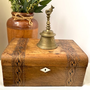 May include: A wooden box with decorative inlaid patterns, a brass bell with a deer-shaped handle, and a brown ceramic vase with greenery. The box has a keyhole with a white diamond-shaped escutcheon.