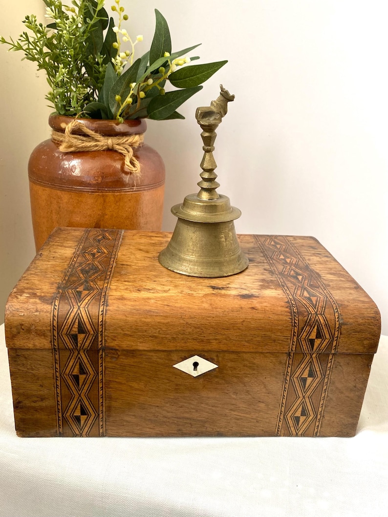 May include: A wooden box with decorative inlaid patterns, a brass bell with a deer-shaped handle, and a brown ceramic vase with greenery. The box has a keyhole with a white diamond-shaped escutcheon.