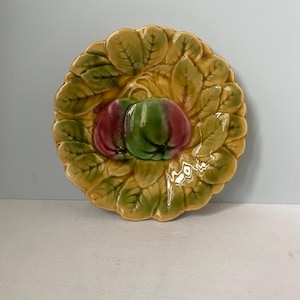 May include: A ceramic plate with a raised design of leaves and fruit. The plate features two fruits, one red and one green, surrounded by a ring of yellow and green leaves. The plate has a glossy finish, suitable for serving food.