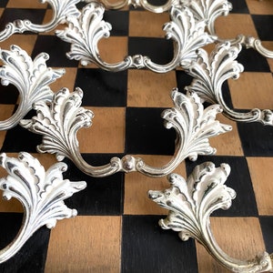 May include: A collection of ornate silver drawer pulls with a white, floral design. The pulls are arranged on a black and brown chequered surface.