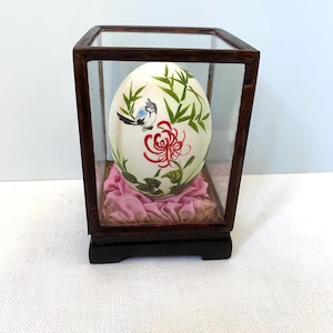 May include: An ornamental egg featuring a painted design of a blue bird, red flower, and green bamboo leaves, presented in a glass and wood display case. The egg sits on a pink fabric cushion within the case, which has a dark brown wooden frame and base.