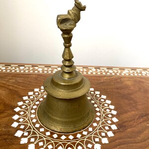 May include: A vintage brass bell with a deer-shaped handle. The bell has a conical shape and sits on a wooden surface with a white floral inlay design. The bell is a warm, antique gold colour.