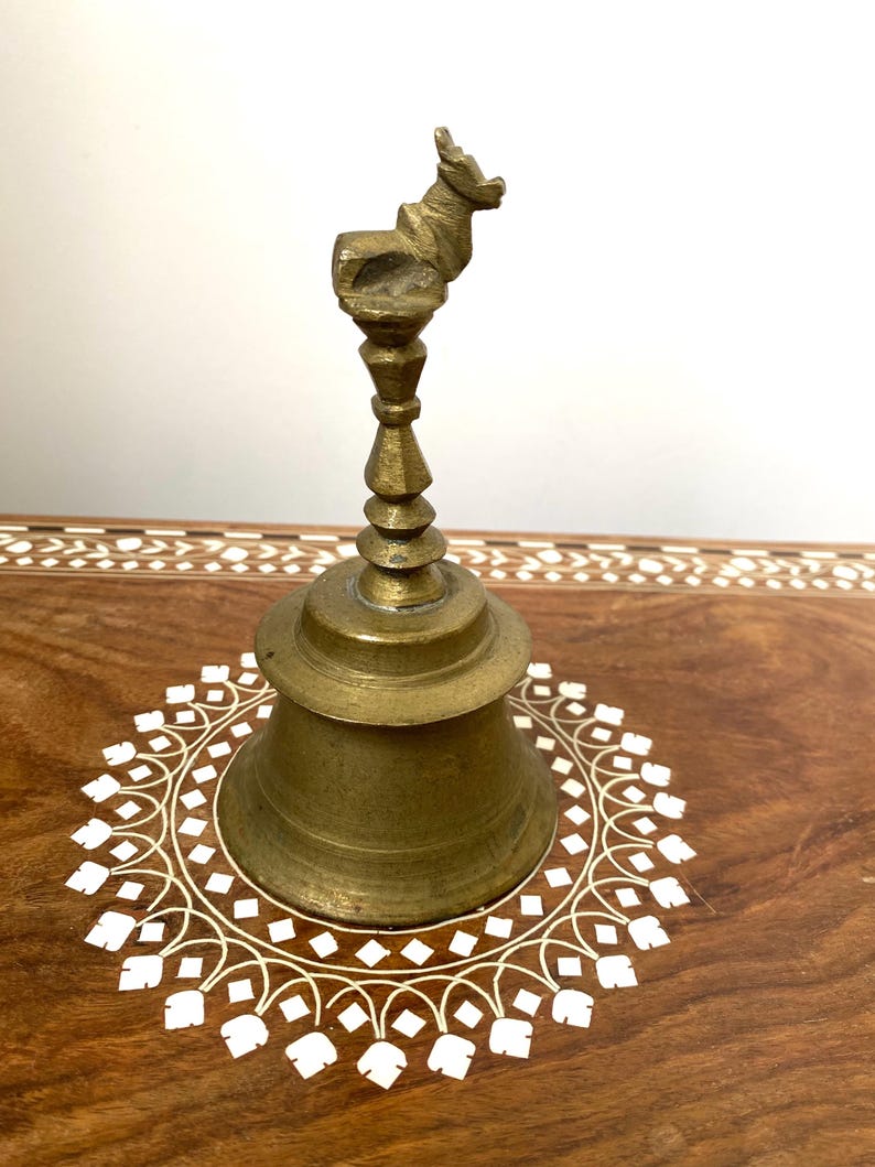 May include: A vintage brass bell with a deer-shaped handle. The bell has a conical shape and sits on a wooden surface with a white floral inlay design. The bell is a warm, antique gold colour.