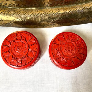 以下が含まれることがあります： 精巧な花の彫刻が施された、2つの丸い鮮やかな赤い箱。箱には、詳細な葉に囲まれた中央のバラのデザインが施されています。背景には、金色の装飾トレイの一部が含まれています。