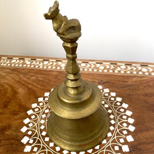 May include: A vintage brass bell with an animal figure on top. The bell has a flared base and an ornate handle. The bell is set on a wooden surface with a white floral design.