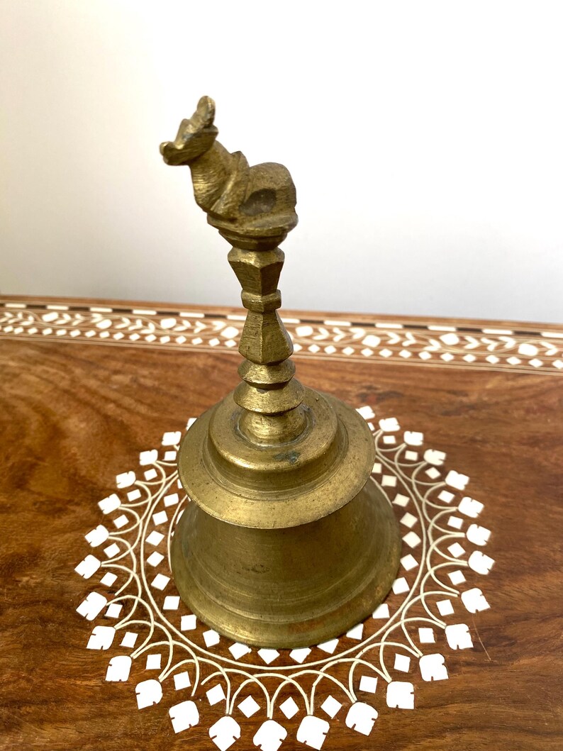 May include: A vintage brass bell with an animal figure on top. The bell has a flared base and an ornate handle. The bell is set on a wooden surface with a white floral design.
