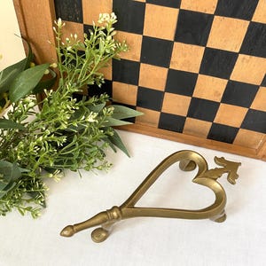 Puede incluir: Un posa ollas de metal dorado en forma de corazón con un diseño decorativo. El posa ollas está sobre una superficie blanca.