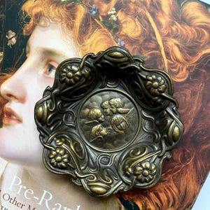 Puede incluir: Un plato decorativo de bronce con motivos florales y de querubines. El plato está colocado sobre un libro titulado "Pre-Raphaelite Other Masters". El plato tiene un diseño ornamentado con un relieve central de querubines.