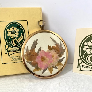 以下が含まれることがあります： クリーム色の背景に、ピンクと茶色の花と緑の葉が特徴の、丸い金色の押し花ペンダント。ペンダントの隣には、緑色の花のロゴと「Ravenwood Crafts」の文字が入った箱とカードがあります。