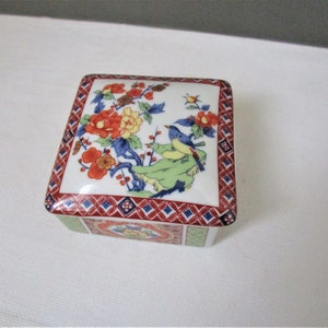 May include: Square porcelain trinket box with a floral and bird design. The box has a red, blue and green geometric pattern around the edge.