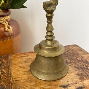 May include: A vintage brass bell with an animal figure on top. The bell has a flared base and a detailed stem. The bell is set on a wooden surface with a decorative pattern. The bell is likely used for decorative purposes. The bell's height is approximately 15 cm.