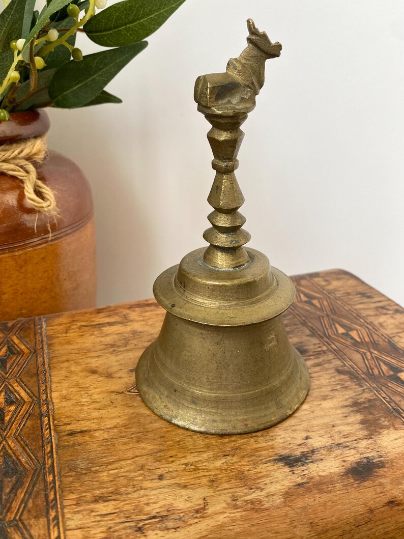 May include: A vintage brass bell with an animal figure on top. The bell has a flared base and a detailed stem. The bell is set on a wooden surface with a decorative pattern. The bell is likely used for decorative purposes. The bell's height is approximately 15 cm.