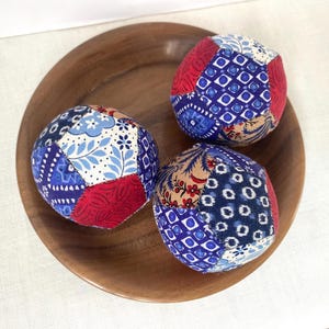 Puede incluir: Tres bolas decorativas de tela con patrones de patchwork en rojo, azul y blanco, dispuestas sobre un plato de madera. Las bolas tienen forma esférica y están hechas de varias telas estampadas.