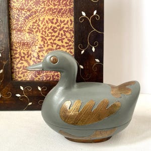 May include: A gray duck-shaped trinket box with gold-colored feather designs. The duck has a copper-colored eye and beak. A decorative wooden frame with a floral design is in the background.