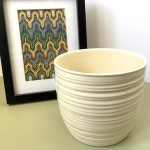 Puede incluir: Jardinera de cerámica color crema con detalles de estrías horizontales. En el fondo, una obra de arte enmarcada con un patrón geométrico en amarillo, gris y verde. La jardinera está sobre una superficie verde claro.