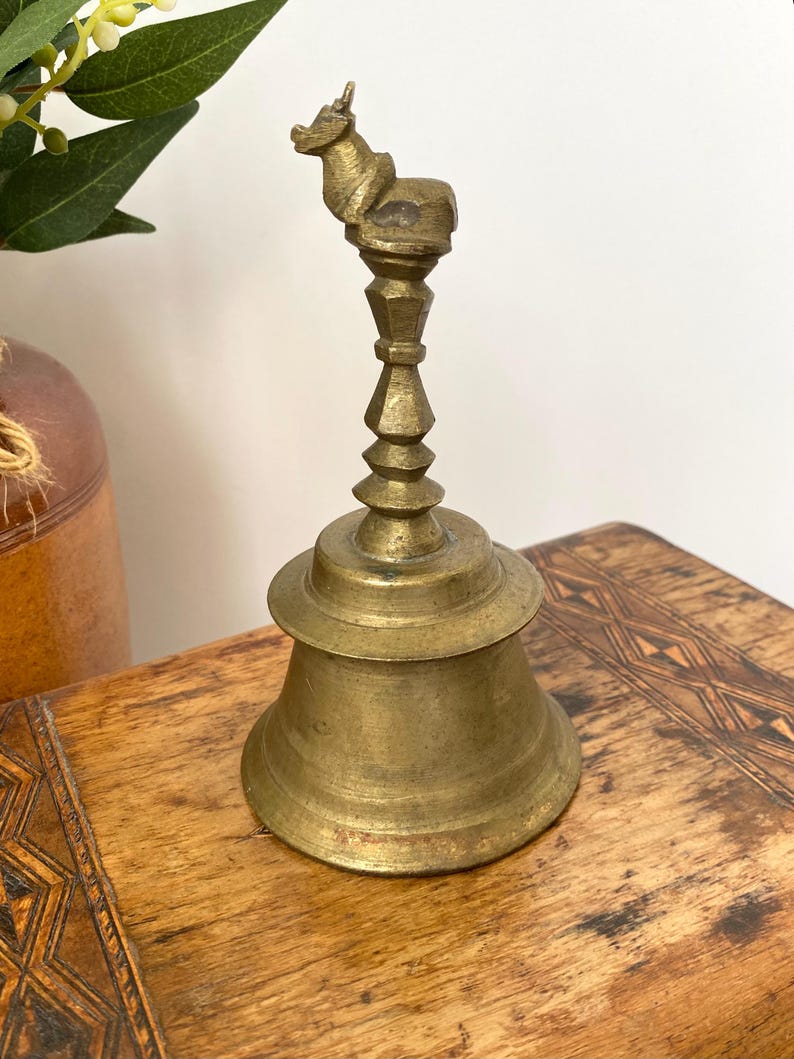 May include: A vintage brass bell with a decorative handle featuring a small animal figure. The bell has a flared base and a detailed, tiered stem. The bell is displayed on a wooden surface.