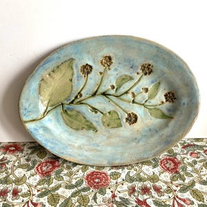 May include: Oval ceramic serving dish with a light blue glaze. The dish features a raised floral design with leaves and small flower clusters in shades of green and brown. The dish is approximately 30 cm long.