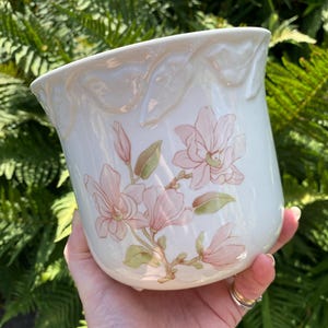 Puede incluir: Maceta de cerámica color crema con diseño floral. La maceta presenta un diseño de flor de magnolia en tonos rosa y verde. El borde superior tiene un diseño ondulado decorativo. La maceta mide unos 15 cm de alto.