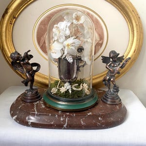 Puede incluir: Una exhibición decorativa con una campana de cristal que muestra flores blancas en un jarrón plateado, sobre una base de mármol. Dos figuras de querubines de bronce flanquean la campana. Una obra de arte ovalada con marco dorado en el fondo.