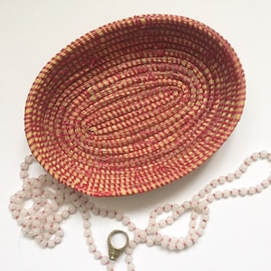 Puede incluir: Una cesta ovalada tejida con un patrón de paja natural y roja. La cesta está vacía y se encuentra sobre una superficie blanca. Un collar de cuentas blancas con un anillo de oro está al lado de la cesta.