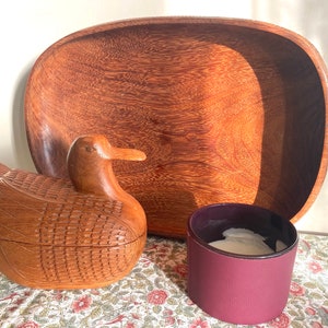 May include: A wooden duck shaped box with a carved design sits next to a wooden serving tray and a burgundy candle holder.