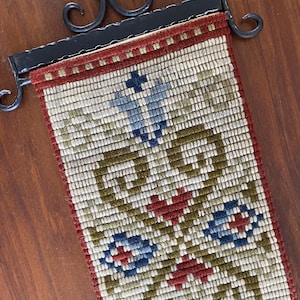 May include: A rectangular tapestry with a cream background and a repeating pattern of blue, red, and green floral motifs. The tapestry is framed with a red border and is suspended from a black wrought iron hanger.