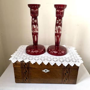 May include: A pair of ruby red glass candlesticks with white floral patterns. They sit on a wooden box with inlaid decoration, covered by a white lace doily. The candlesticks have a classic, elegant design.