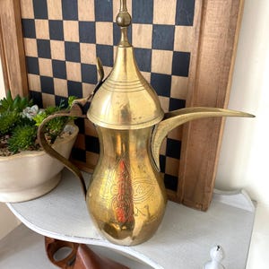 Puede incluir: Una cafetera de latón vintage con un pico largo y detalles ornamentados. La cafetera tiene una tapa cónica y un asa. El fondo incluye un tablero de ajedrez enmarcado en madera y un estante blanco.