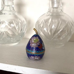 May include: A small, pear-shaped trinket box with a blue enamel finish, decorated with floral and geometric patterns in pink, green, and gold. The box has a gold-coloured stem and a turquoise border. Two clear glass bottles are in the background.