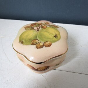 Puede incluir: Pequeña caja de baratijas decorativa con base y tapa de color rosa pálido. La tapa presenta un diseño pintado de dos membrillos verdes, nueces marrones y ribetes dorados. La caja tiene una forma redondeada única.