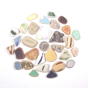 May include: A collection of assorted smooth sea glass and ceramic shards in various shapes and colors, including white, blue, yellow, brown, and green pieces, arranged around a single silver-colored United States dime for scale reference.